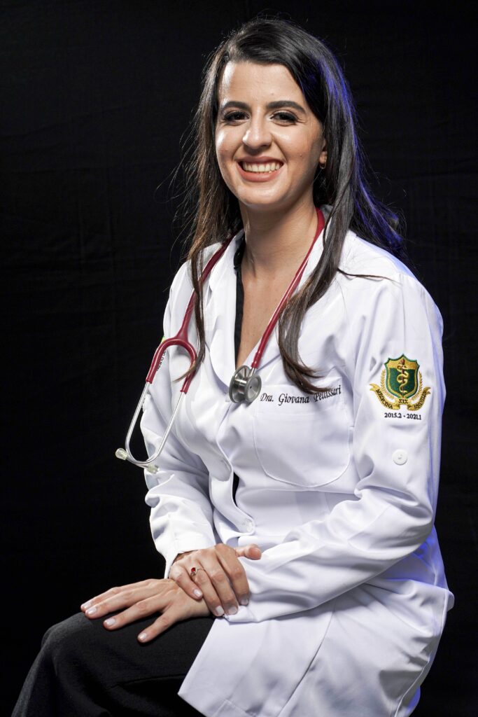 Dra Giovana de jaleco branco, com um estetoscópio vermelho em seus ombros, sentada com as mãos cruzadas sobre a perna.