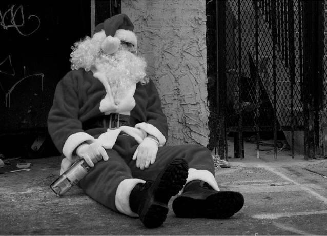 Imagem em preto e branco com homem vestido de papai noel, sentado no chão da rua com uma garrafa de bebida alcoolica na mão.