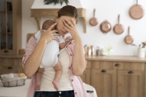Mulher segurando um bebê recém nascido, com uma mão sobre a testa, representando a depressão pós-parto