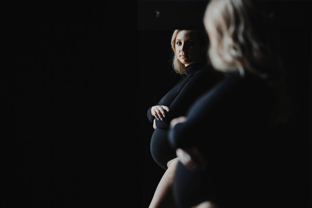 Mulher grávida com as mãos sobre a barriga e olhar triste, se olhando no espelho, com a cor predominante preta na roupa e no fundo, representando a depressão na gestação