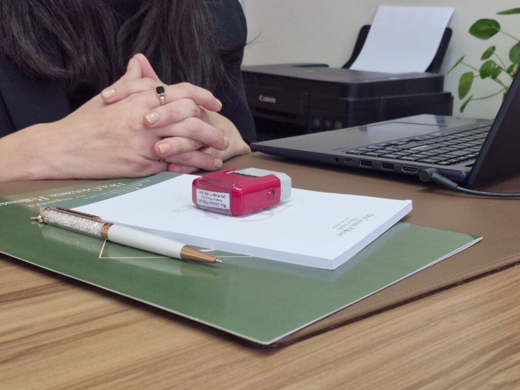 A imagem mostra as mãos da dra Giovana sobre sua mesa em seu consultório, com pasta, receituário, carimbo e caneta sobre a mesa, um notebook ao lado.