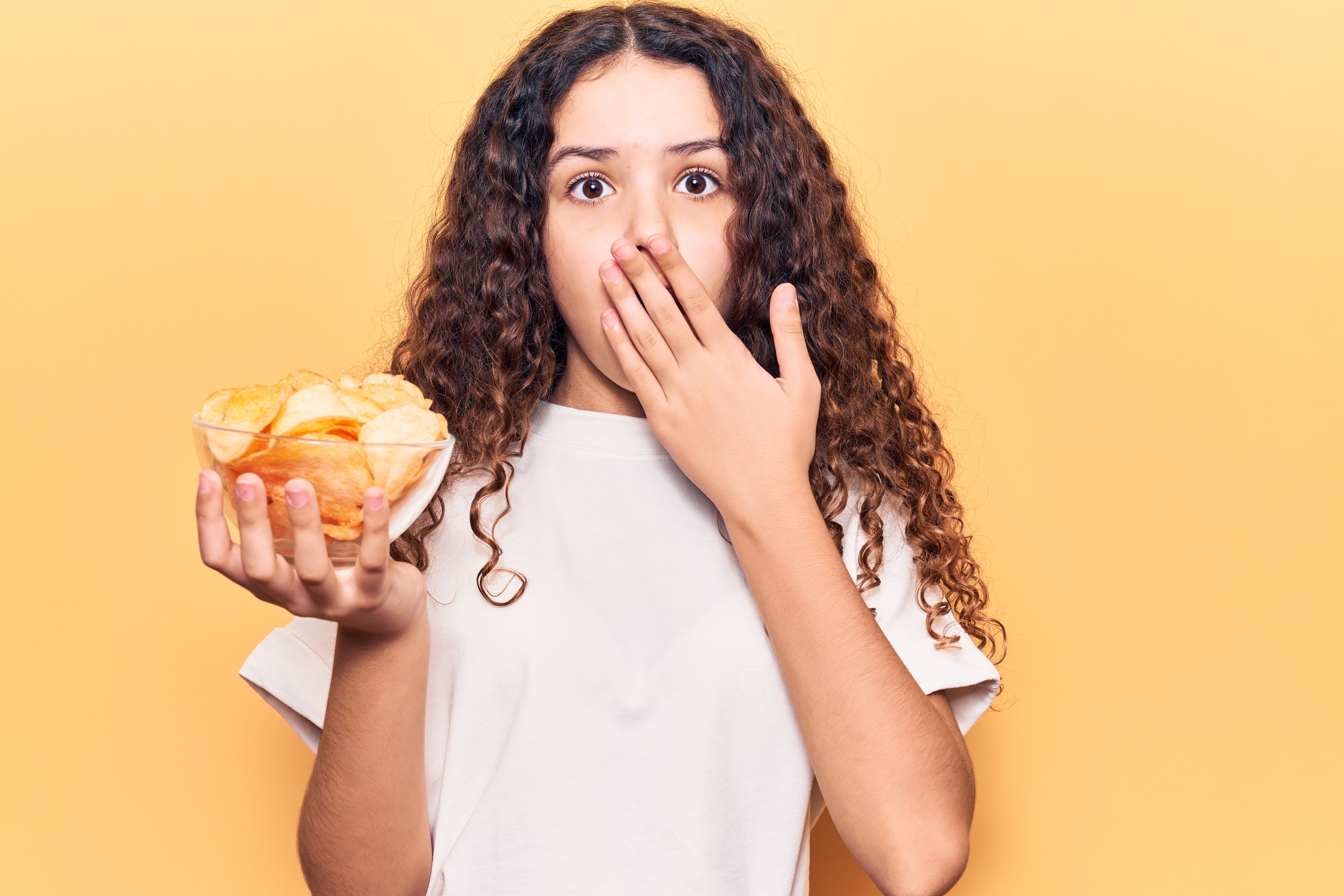 Menina adolescente de cabelos cacheados e camiseta branca, segurando um pote transparente com batatas chips dentro, e com a outra mão na boca, e expressão de susto ou medo.