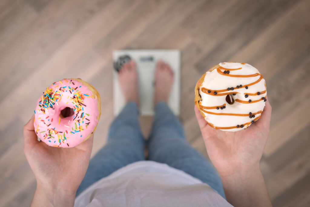 A imagem demonstra o enquadramento de uma pessoa olhando para baixo, vendo seus pés sobre uma balança, segurando um donuts em cada uma das mãos.