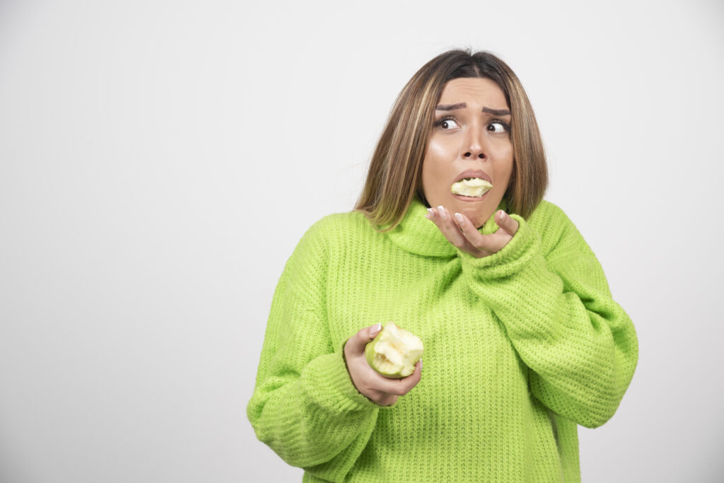 Mulher de roupa verde, segurando uma maça verde na mão,com a boca cheia de maçã e cara de desconforto. A outra mão esta perto da boca, para segurar a maça caso caia da boca. Representa os transtornos alimentares.