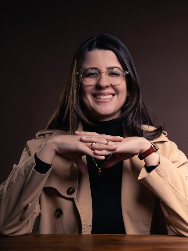 Dra Giovana, sentada, com os cotovelos sobre uma mesa e mãos entrecruzadas à sua frente, com um grande sorriso.