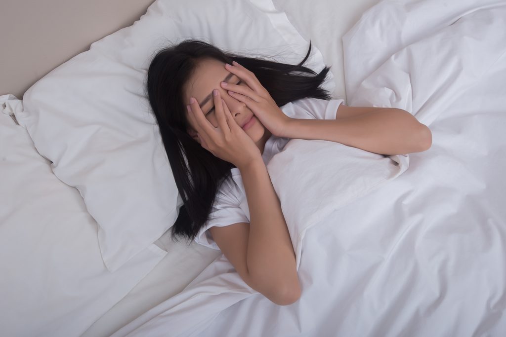 Foto de uma mulher deitada em uma cama com roupas de cama brancas e confortáveis, mas com as mãos sobre o rosto e expressão de cansaço, representando a insônia.